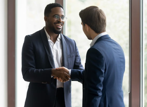 Two men shaking hands next to a wall of windows with trees outside.